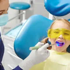 Dziecko w okularach ochronnych u dentysty. Lekarz utwardza lak światłem. Bez obaw o lakowanie zębów skutki uboczne.