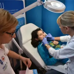 Dziecko na fotelu stomatologicznym, badane przez lekarkę. Na plakacie w tle widnieje napis "ŻUJ" i logo Luxmed. Sprawdź luxmed stomatologia cennik.