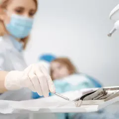Dentystka w masce przygotowuje narzędzia do ekstrakcji zęba. W tle pacjentka na fotelu.