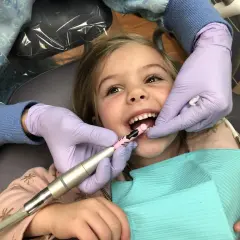 Uśmiechnięta dziewczynka przechodzi zabieg lapisowania zębów. Dentysta w rękawiczkach delikatnie czyści jej ząbki.