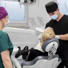 Dentysta w masce i czarnym uniformie wykonuje zabieg. Obok stoi asystentka w zielonym fartuchu. To zdjęcie szwów po zabiegu.