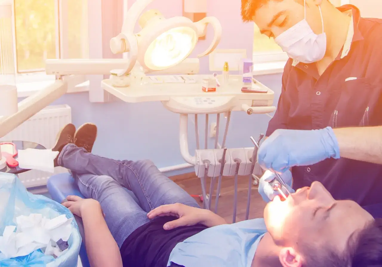 Dentysta w masce i rękawiczkach przeprowadza ekstrakcję zęba u pacjenta leżącego na fotelu.
