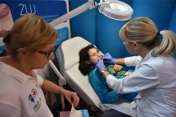 Dziecko na fotelu stomatologicznym, badane przez lekarkę. Na plakacie w tle widnieje napis "ŻUJ" i logo Luxmed. Sprawdź luxmed stomatologia cennik.