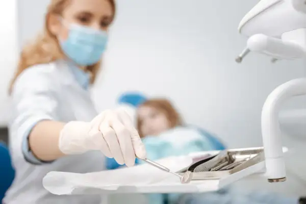 Dentystka w masce przygotowuje narzędzia do ekstrakcji zęba. W tle pacjentka na fotelu.