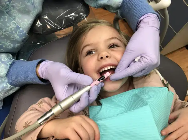 Uśmiechnięta dziewczynka przechodzi zabieg lapisowania zębów. Dentysta w rękawiczkach delikatnie czyści jej ząbki.